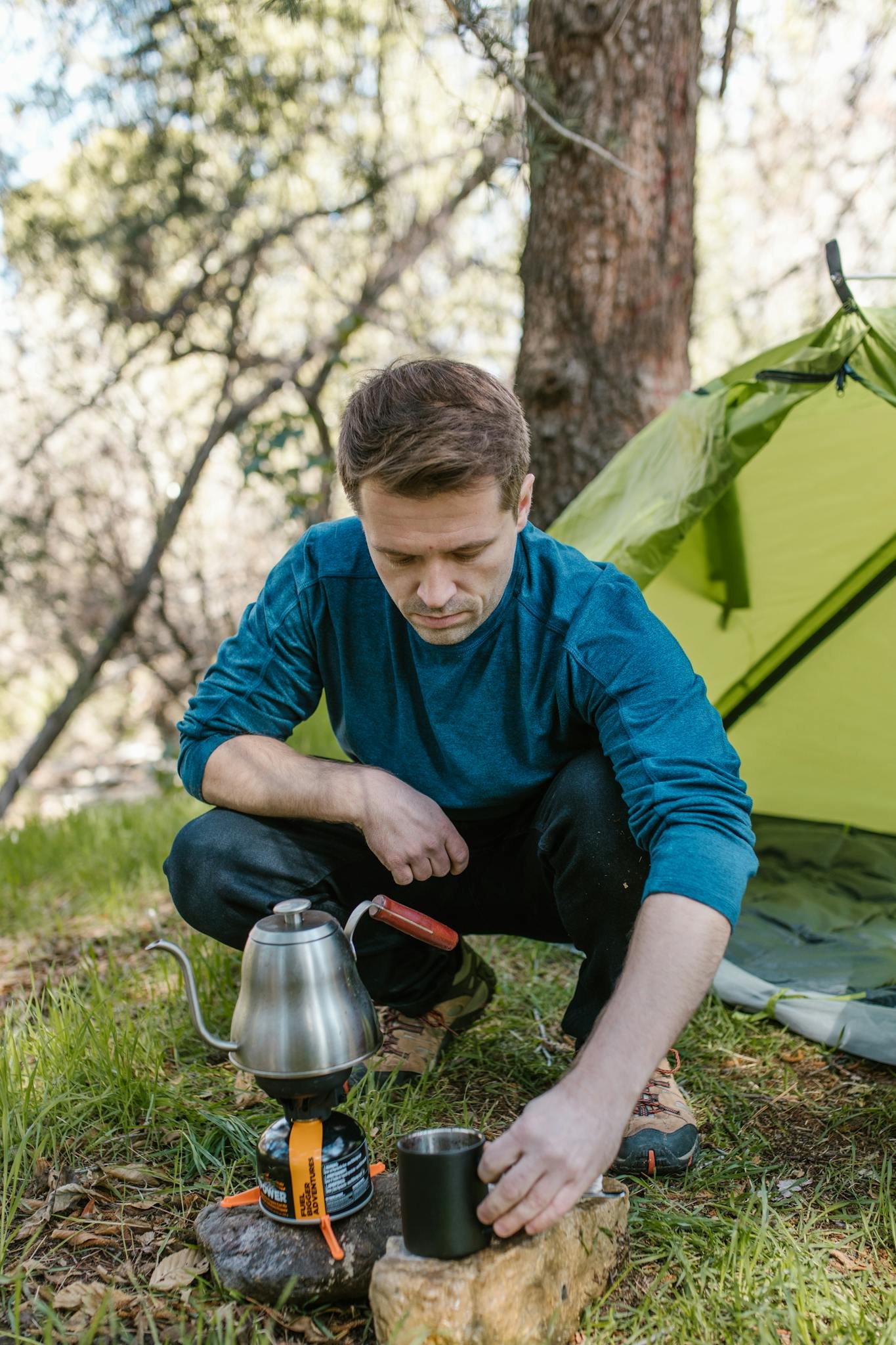 A man prepares coffee at a campsite using a portable gas burner and kettle.
