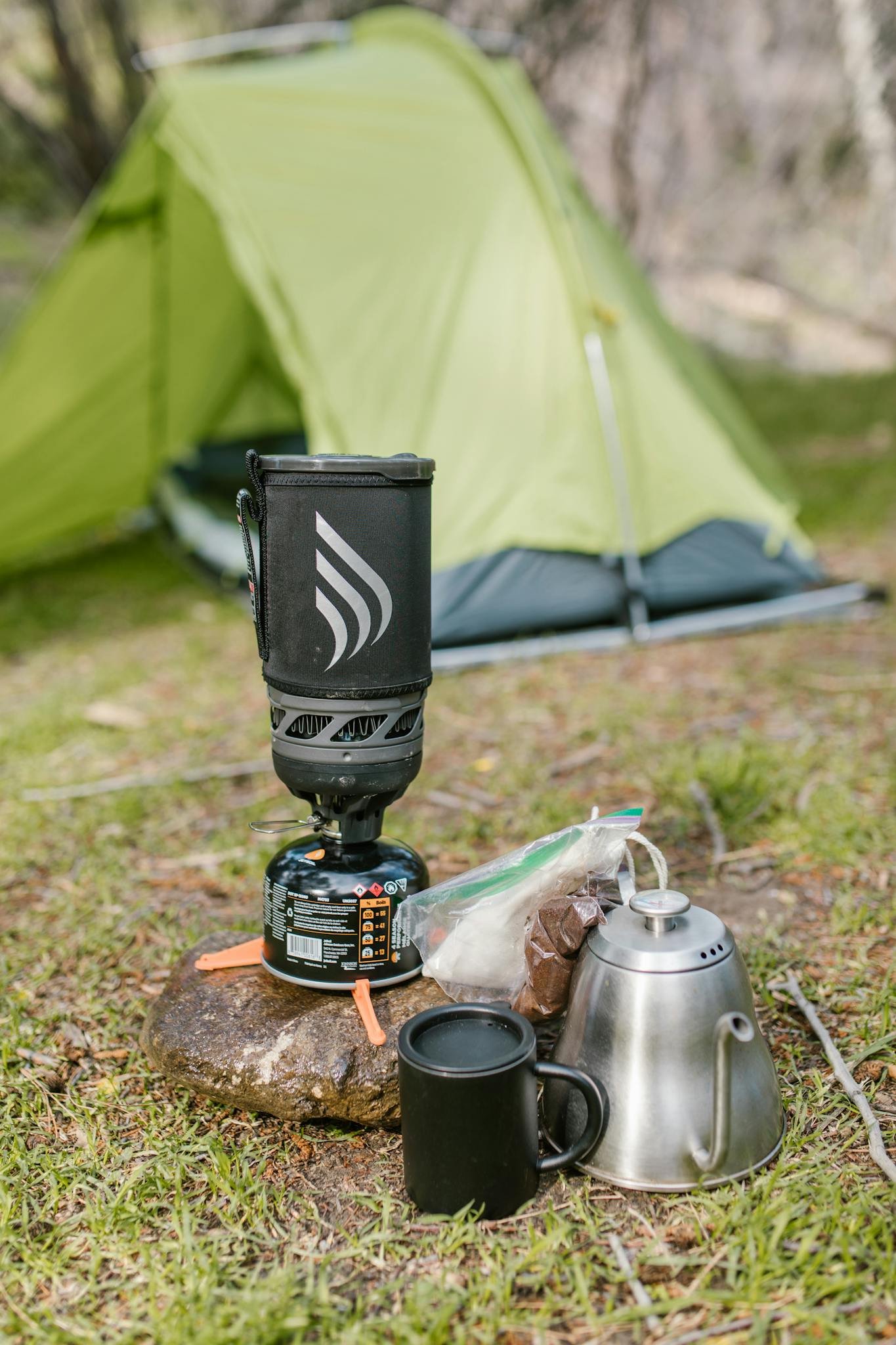 Camp setup with coffee tools, portable stove, and green tent in a natural setting.